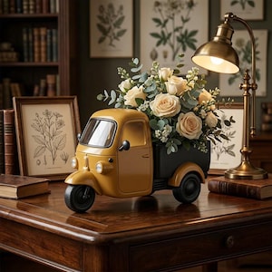 Può includere: Una statuetta di camion a tre ruote color giallo senape con un cassone nero riempito da un bouquet di rose color crema e foglie di eucalipto verdi. Il veicolo in miniatura presenta un faro anteriore, un parabrezza e ruote nere.