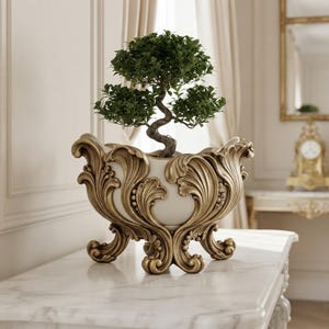 Puede incluir: Un pequeño bonsái verde plantado en una maceta blanca con forma de cuenco. La maceta presenta un exterior ornamentado en tono dorado metálico con intrincados relieves de hojas de acanto y cuatro patas curvas decorativas en la base.