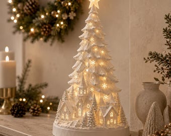 Puede incluir: Una escultura de árbol de Navidad blanca, cónica e iluminada, con una estrella en la punta, recortes de copos de nieve y una escena de pueblo en miniatura en la base. La estructura se asienta sobre un pedestal circular blanco y emite luz.