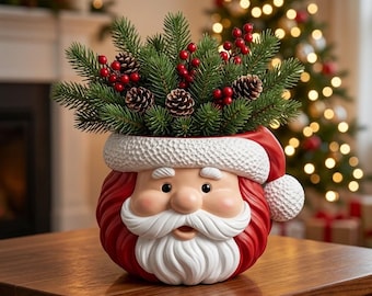 Puede incluir: Una maceta de cerámica con forma de Santa Claus que presenta un gorro rojo con borde texturizado blanco y pompón, bigote y barba blancos. La maceta está llena de ramas de pino verdes, pequeñas piñas marrones y bayas rojas brillantes.
