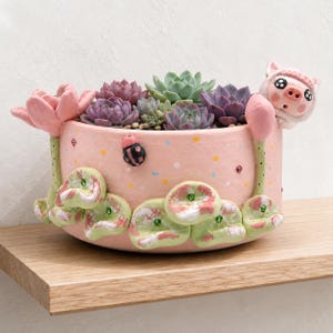 May include: A round, light pink ceramic planter filled with various green and purple succulents. The exterior features sculpted pink lotus flowers, green lily pads with small green rhinestones, a black ladybug, and a small pig figure.
