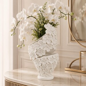 May include: A white ceramic vase featuring a spiraling, layered design covered in sculpted floral reliefs. The vase holds a large arrangement of white orchid stems with green buds and foliage, creating a textured, monochromatic display.