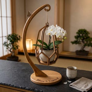 May include: A wooden stand with a curved arm and a circular base featuring the text "Bloom & Inspire" holds a hanging geometric wooden frame containing a white ceramic pot with a white orchid plant. A small wooden butterfly sits on the arm.