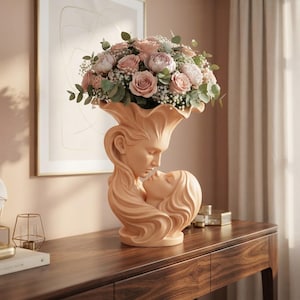 Pode incluir: Um vaso de cerâmica cor de pêssego em forma de dois rostos num abraço, com cabelos fluidos formando a base e a borda. O vaso está cheio com um bouquet de rosas cor-de-rosa claro, gipsófila branca e folhas de eucalipto verdes.