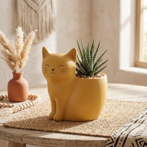 Op de afbeelding: Een mosterdgele keramische plantenbak in de vorm van een zittende kat met een gladde afwerking. De kat heeft eenvoudige zwarte lijnen voor de ogen, neus en snorharen. Een groen-wit gestreepte vetplant staat in de rug van de kat geplant.