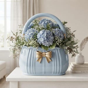 May include: A light blue ceramic basket-shaped vase with a vertical ribbed texture and a central gold-coloured bow accent. The vase is filled with a lush arrangement of light blue hydrangea blossoms, small white baby's breath, and green foliage.