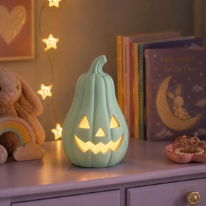 Op de afbeelding: Een licht mintgroene pompoenvormige lamp met een uitgesneden jack-o'-lantern gezicht dat van binnenuit oplicht. De lamp heeft een hoog, geribbeld ontwerp en staat op een oppervlak, waarbij warm, zacht licht door de ogen en mond schijnt.