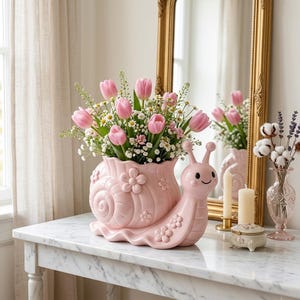 May include: A light pink ceramic snail-shaped planter featuring a shell decorated with raised flower motifs. The planter is filled with a bouquet of pink tulips and small white flowers. The snail has a smooth finish and a simple smiling face.