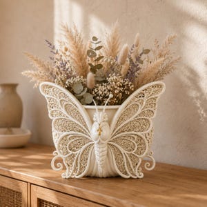 May include: A cream-colored butterfly-shaped vase featuring intricate, lace-like filigree patterns on its wings. The vase is filled with a dried floral arrangement consisting of pampas grass, eucalyptus leaves, lavender, and small white flowers.