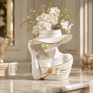 May include: A white bust-shaped vase featuring a figure wearing a wide-brimmed hat with gold trim and a bow detail on the bodice. The figure is holding the hat brim with one hand and is filled with white orchid flowers and green stems.