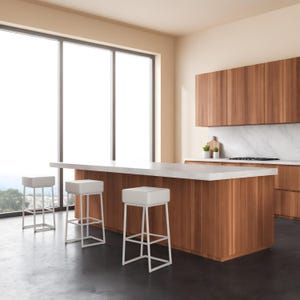 Könnte beinhalten: Drei weiße Barhocker mit quadratischen Sitzen und dünnen weißen Metallrahmen vor einer Kücheninsel mit weißer Arbeitsplatte und Holzschränken. Die Küche verfügt über einen Marmor-Spritzschutz und große Fenster mit natürlichem Licht.