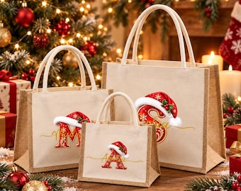 Puede incluir: Tres bolsas de yute beige de diferentes tamaños, cada una con una letra roja con detalles de filigrana dorada rematada por un gorro de Papá Noel rojo con borde blanco. Las bolsas tienen asas resistentes y una textura tejida natural.