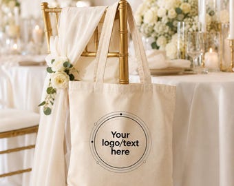 Può includere: Una borsa tote in tela beige con due tracolle, caratterizzata da un design grafico circolare nero sul davanti con il testo "Your logo/text here" centrato all'interno del cerchio. La borsa è appesa a una sedia in metallo color oro.