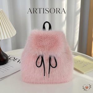 May include: A light pink faux fur backpack featuring a black top handle and black drawstring cords with silver metal tips. The word ARTISORA is printed in black above the bag. The backpack has a soft, textured appearance and a flap closure.
