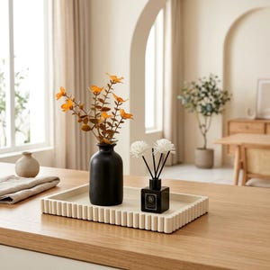 May include: A rectangular off-white tray with a fluted edge holds a matte black vase containing orange artificial flowers and a small black square glass bottle with a reed diffuser featuring two white flower-shaped reeds and black sticks.
