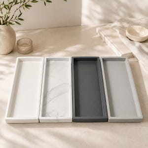 May include: Four rectangular trays arranged side by side. The first tray is solid white, the second features a white and gray marble pattern, the third is dark charcoal gray, and the fourth is a light gray. Each tray has a flat base and raised edges.