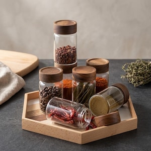 Puede incluir: Un juego de seis frascos de vidrio transparente con tapas de madera marrón oscuro, dispuestos sobre una bandeja hexagonal de madera clara. Los frascos contienen varias especias, granos de café y chiles secos, un frasco está acostado.