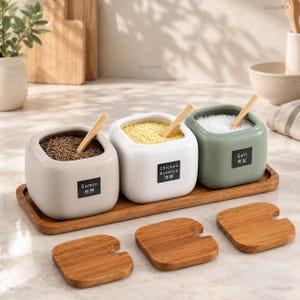 Puede incluir: Tres tarros cuadrados de cerámica para especias en gris claro, blanco y verde salvia, cada uno con una cuchara de madera y una etiqueta negra que dice Sumin, Chicken Essence o Salt. Los tarros descansan sobre una bandeja de madera con tres tapas.