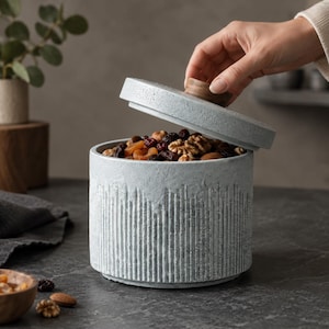 Puede incluir: Un tarro de almacenamiento cilíndrico de cerámica gris claro con un exterior texturizado de rayas verticales y una tapa a juego con un pequeño pomo de madera. El tarro está lleno de una variedad de frutos secos y frutas deshidratadas.