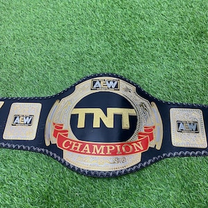 May include: A black leather championship wrestling belt featuring a large central plate with the gold text TNT and a red banner reading CHAMPION. The belt includes the All Elite Wrestling logo on the center and side plates with gold accents.