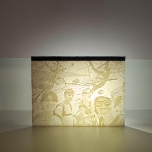 Peut inclure: Une lampe lithophane rectangulaire imprimée en 3D présentant une image en relief monochrome de quatre personnes debout à l'extérieur avec des arbres en arrière-plan. La source lumineuse interne illumine le matériau translucide.