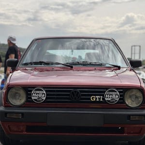 Peut inclure: Une Volkswagen Golf GTI bordeaux vue de face. Le véhicule présente une calandre noire avec un emblème Volkswagen central, deux phares ronds et deux feux auxiliaires circulaires Hella. Le badge GTI est visible sur la calandre avant.
