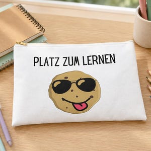 Könnte beinhalten: Ein weißes, rechteckiges Federmäppchen mit Reißverschluss, das den schwarzen Schriftzug "PLATZ ZUM LERNEN" über einer Grafik eines beigen Kekses mit schwarzer Sonnenbrille, lächelndem Mund und herausgestreckter rosa Zunge zeigt.