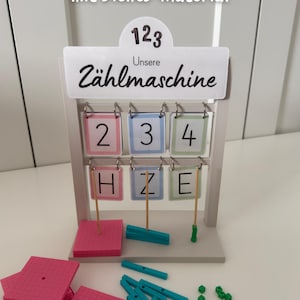 Könnte beinhalten: Eine hellgraue hölzerne Zählmaschine mit einem Schild mit der Aufschrift "Unsere Zählmaschine" und "123". Sie verfügt über zwei Reihen hängender Karten mit den Zahlen 2, 3, 4 und den Buchstaben H, Z, E. Enthält rosa quadratische Platten, türkisfarbene Stäbe und kleine grüne Würfel.