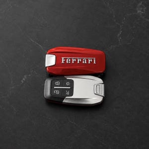 Könnte beinhalten: Ein Ferrari-Autoschlüssel mit einem glänzend roten Gehäuse, das den silbernen Ferrari-Schriftzug auf der oberen Hälfte trägt, sowie einem silber-schwarzen Bedienfeld auf der unteren Hälfte mit vier Tasten für Verriegelung, Entriegelung und Licht.