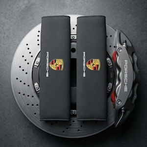 May include: Two rectangular black seat belt covers featuring the Porsche logo and crest in color. These are positioned over a metallic silver perforated brake disc with a grey brake caliper that also displays the Porsche brand name in white text.