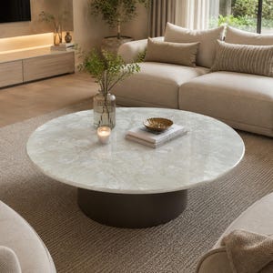Puede incluir: Una mesa de centro redonda con una superficie de piedra blanca pulida con vetas grises sutiles y una base cilíndrica de color marrón oscuro. Sobre la mesa hay un jarrón de vidrio transparente con follaje verde, una vela blanca y dos libros.