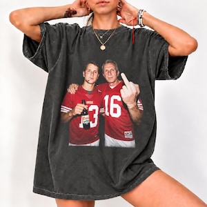 Könnte beinhalten: Ein anthrazitfarbenes Kurzarm-T-Shirt mit einem Grafikdruck von zwei Footballspielern in roten Trikots. Ein Spieler hält eine Bierflasche und hat eine Zigarette im Mund, während der andere Spieler den Mittelfinger in die Höhe streckt.