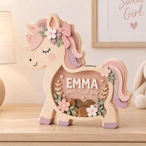 May include: A wooden unicorn-shaped piggy bank with a clear front panel containing coins. The design features pastel pink, purple, and green accents, a floral mane, and the name EMMA printed above the text sparkle, bloom, grow in white.