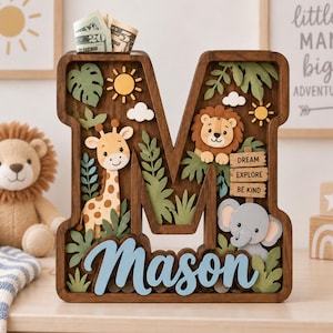 Può includere: Un salvadanaio in legno a forma di lettera M con tema giungla, raffigurante una giraffa, un leone e un elefante. Il design include foglie verdi, soli gialli e nuvole bianche. Un cartello in legno riporta la scritta Dream Explore Be Kind.