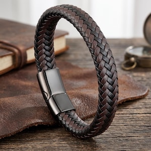 May include: A braided bracelet featuring alternating strands of dark brown and black leather. The bracelet is secured with a smooth, polished gunmetal-coloured rectangular magnetic clasp that connects the two ends of the woven leather band.