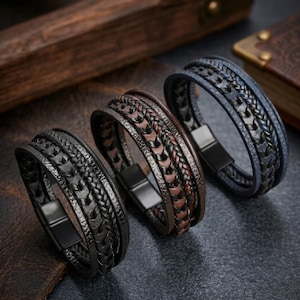 May include: Three multi-strand bracelets featuring braided and textured leather-like cords in black, brown, and blue. Each bracelet is secured with a rectangular black metal magnetic clasp, showcasing a layered design with intricate woven patterns.