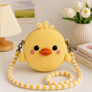 Pode incluir: Uma bolsa tiracolo redonda de crochê amarela em forma de pintinho. Possui olhos de botão pretos, um pequeno bico laranja, bochechas bordadas em rosa e um tufo de penas no topo. A bolsa inclui uma alça de ombro removível com listras amarelas e brancas.
