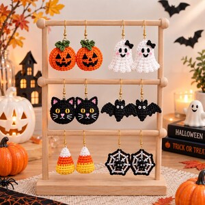 Pode incluir: Seis pares de brincos de crochê feitos à mão para o Halloween pendurados num suporte de madeira. Os designs incluem abóboras cor de laranja, fantasmas brancos, gatos pretos, morcegos, doces e teias de aranha pretas com aranhas, com ganchos dourados.