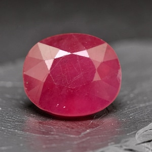 May include: A vibrant, oval-cut ruby gemstone with a deep red hue and multiple triangular facets. The stone displays a polished, reflective surface with visible internal clarity and light refraction across its symmetrical, rounded shape.