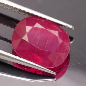 May include: A vibrant, oval-cut ruby gemstone with a deep red hue and multifaceted surface, held securely by a pair of silver-toned metal tweezers. The gemstone displays internal clarity and light reflection across its polished, geometric facets.