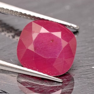 May include: A cushion-cut, vibrant pinkish-red gemstone held by silver-toned metal tweezers. The stone features a faceted surface with a polished finish, displaying a deep, saturated color throughout its structure and a distinct geometric pattern.
