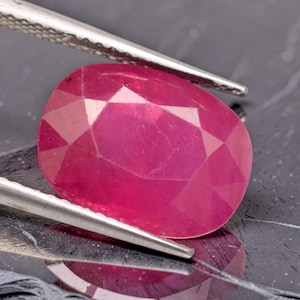 May include: A vibrant, oval-cut, translucent pinkish-red gemstone held by silver-toned metal tweezers. The stone features a faceted surface that reflects light, showcasing its deep color and internal clarity against a dark, reflective surface.