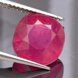 May include: A cushion-cut ruby gemstone with a vibrant pinkish-red hue and multiple facets. The gemstone is held by a pair of silver-toned metal tweezers, showcasing its translucent quality and internal inclusions visible through the polished surface.