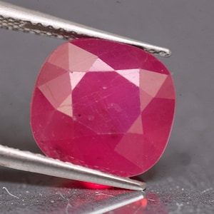 May include: A vibrant, cushion-cut ruby gemstone with a deep pinkish-red hue and multiple triangular and trapezoidal facets. The gemstone is held securely by a pair of silver-toned metal tweezers, showcasing its polished surface and clarity.