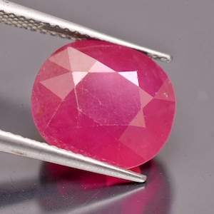 May include: A vibrant, oval-cut ruby gemstone with a deep pinkish-red hue and a faceted surface. The gemstone is held by a pair of silver-toned metal tweezers, showcasing its translucent quality and geometric cut pattern against a neutral background.