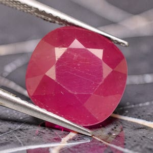 May include: A cushion-cut ruby gemstone with a vibrant deep pinkish-red hue and multifaceted surface. The gemstone is held by a pair of silver-toned metal tweezers, showcasing its transparent clarity and geometric cut against a dark background.