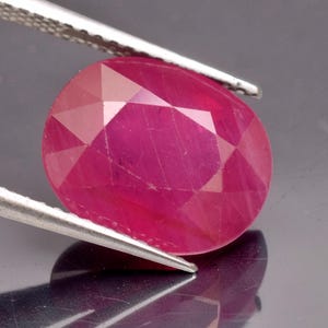 May include: A vibrant, oval-cut ruby gemstone with a deep pinkish-red hue and a faceted surface. The gemstone is held by a pair of silver-toned metal tweezers, showcasing its translucent quality and geometric cut against a dark background.