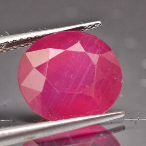 May include: A vibrant, oval-cut ruby gemstone with a deep pinkish-red hue and multiple facets that reflect light. The gemstone is held securely between the two metal tips of a pair of tweezers, showcasing its translucent clarity and polished surface.