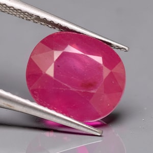 May include: A vibrant, oval-cut ruby gemstone with a deep pinkish-red hue and multifaceted surface, held securely by a pair of silver-toned metal tweezers. The gemstone displays internal clarity and light reflection across its polished facets.