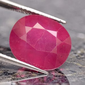 May include: A vibrant, oval-cut ruby gemstone with a faceted surface that reflects light. The deep pinkish-red stone is held by silver-toned metal tweezers, showcasing its translucent quality and internal structure against a dark, textured surface.